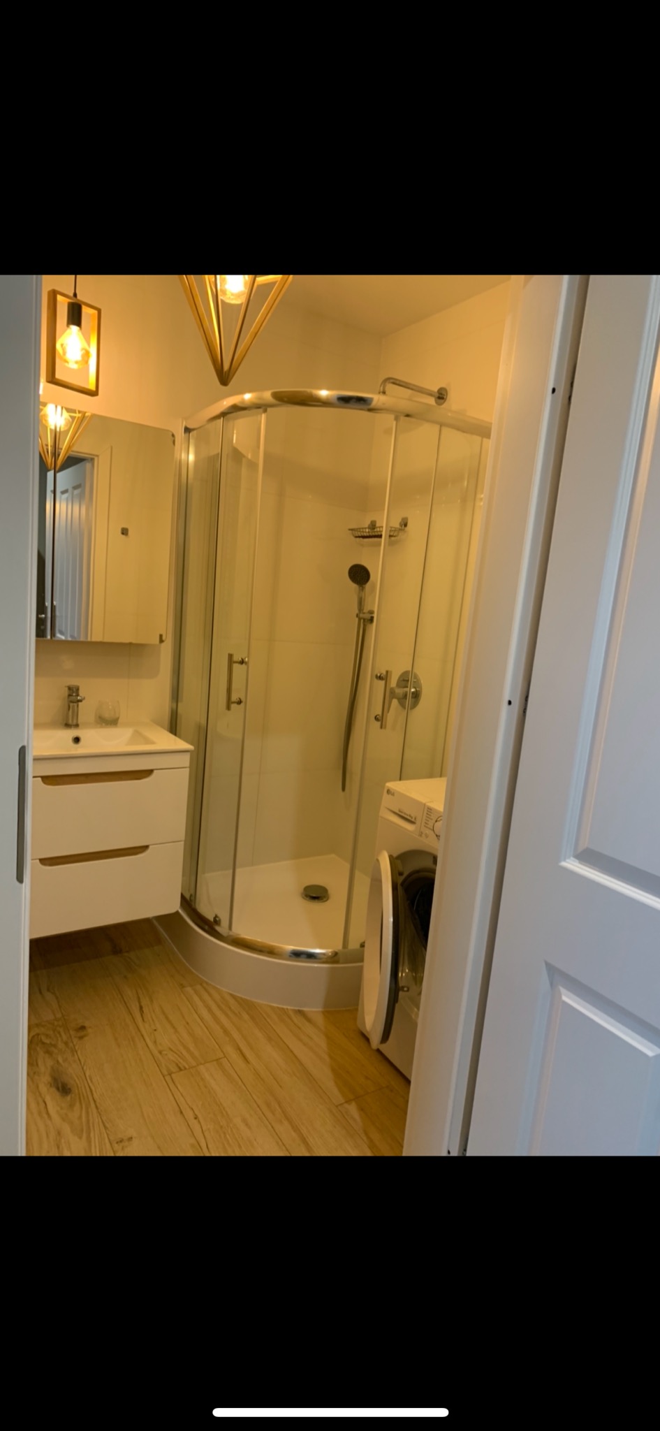 Nowoczesna łazienka z kabiną prysznicową narożną, białą umywalką z szafką, drewnopodobną podłogą i pralką z otwartymi drzwiami, oświetlona lampami w geometrycznych oprawach.