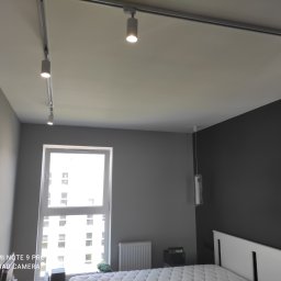 Adwantbud - Szara sypialnia z białym łóżkiem i oknem, oświetlona systemem szynowym z reflektorami punktowymi i lampami wiszącymi w folii ochronnej.