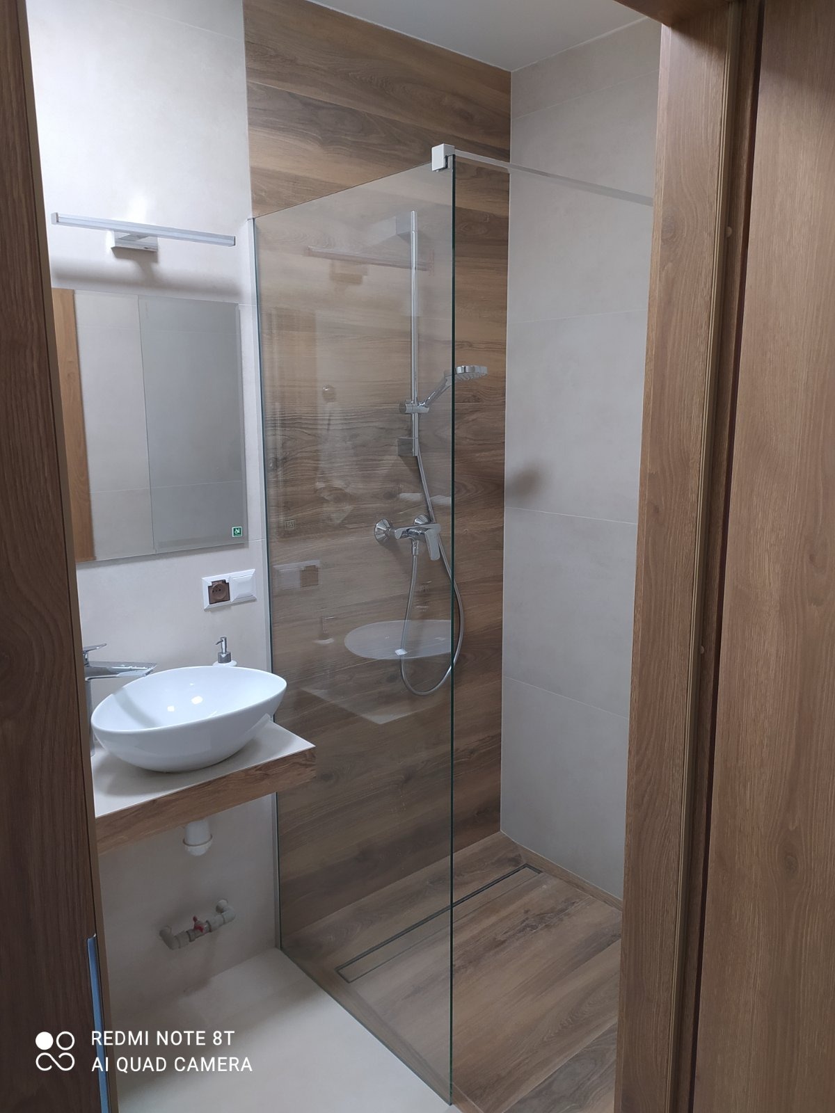 Nowoczesna łazienka z kabiną prysznicową typu walk-in, umywalką nablatową na drewnianym blacie i płytkami imitującymi drewno na ścianach prysznica.