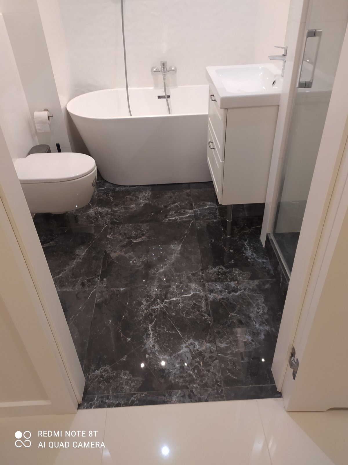 Nowoczesna łazienka z wolnostojącą wanną, toaletą i białą szafką umywalkową na tle ciemnej, marmurkowej podłogi, widok zza progu.