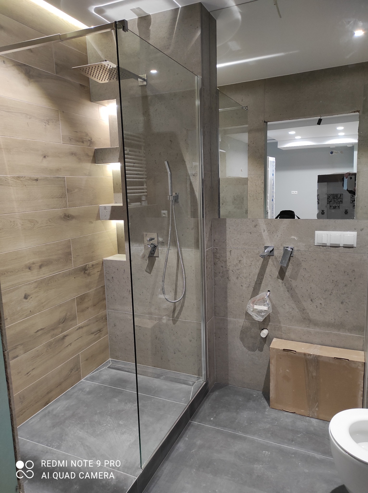 Nowoczesna kabina prysznicowa walk-in z deszczownicą, wykończona płytkami imitującymi drewno na jednej ścianie i szarym kamieniem na pozostałych, z wbudowanym oświetleniem LED w półkach, widoczne...