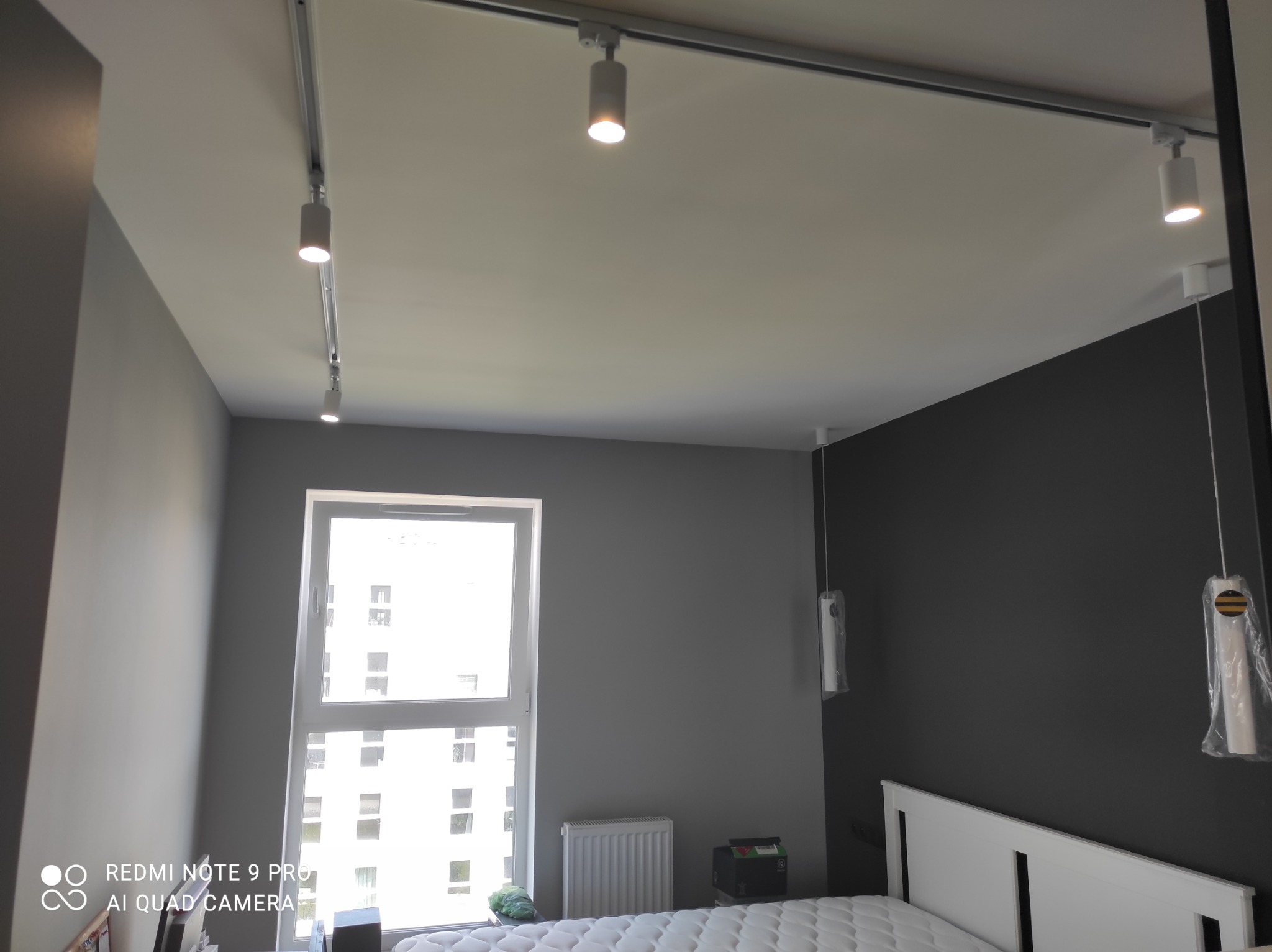 Szara sypialnia z białym łóżkiem i oknem, oświetlona systemem szynowym z reflektorami punktowymi i lampami wiszącymi w folii ochronnej.