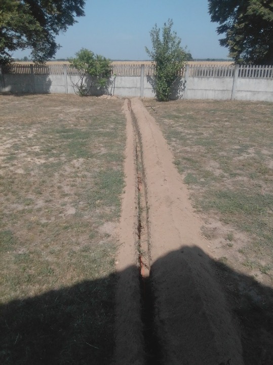 Wykopany wąski rów w ziemi na trawiastym terenie, prowadzący w kierunku betonowego ogrodzenia z drewnianymi elementami, widoczne drzewa i krzewy w tle.