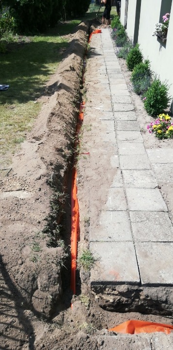 Wykopany rów wzdłuż ścieżki z kostki brukowej, wewnątrz pomarańczowe rury, widoczna trawa, krzewy i fragment budynku z kwiatami w oknie.