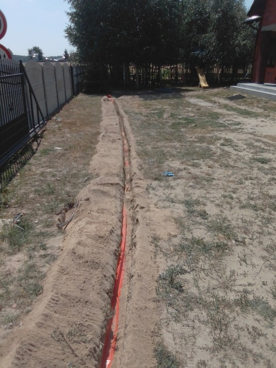 Wykopany rów z pomarańczową rurą w ziemi, widok z góry, obok ogrodzenie i drzewa.