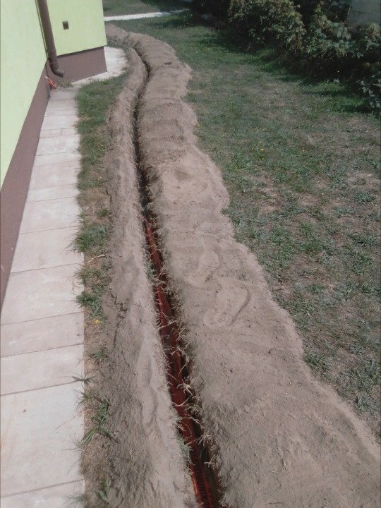 Wykonany wykop pod kable elektryczne z pomarańczową rurą karbowaną w ziemi, widok z góry wzdłuż ściany budynku.