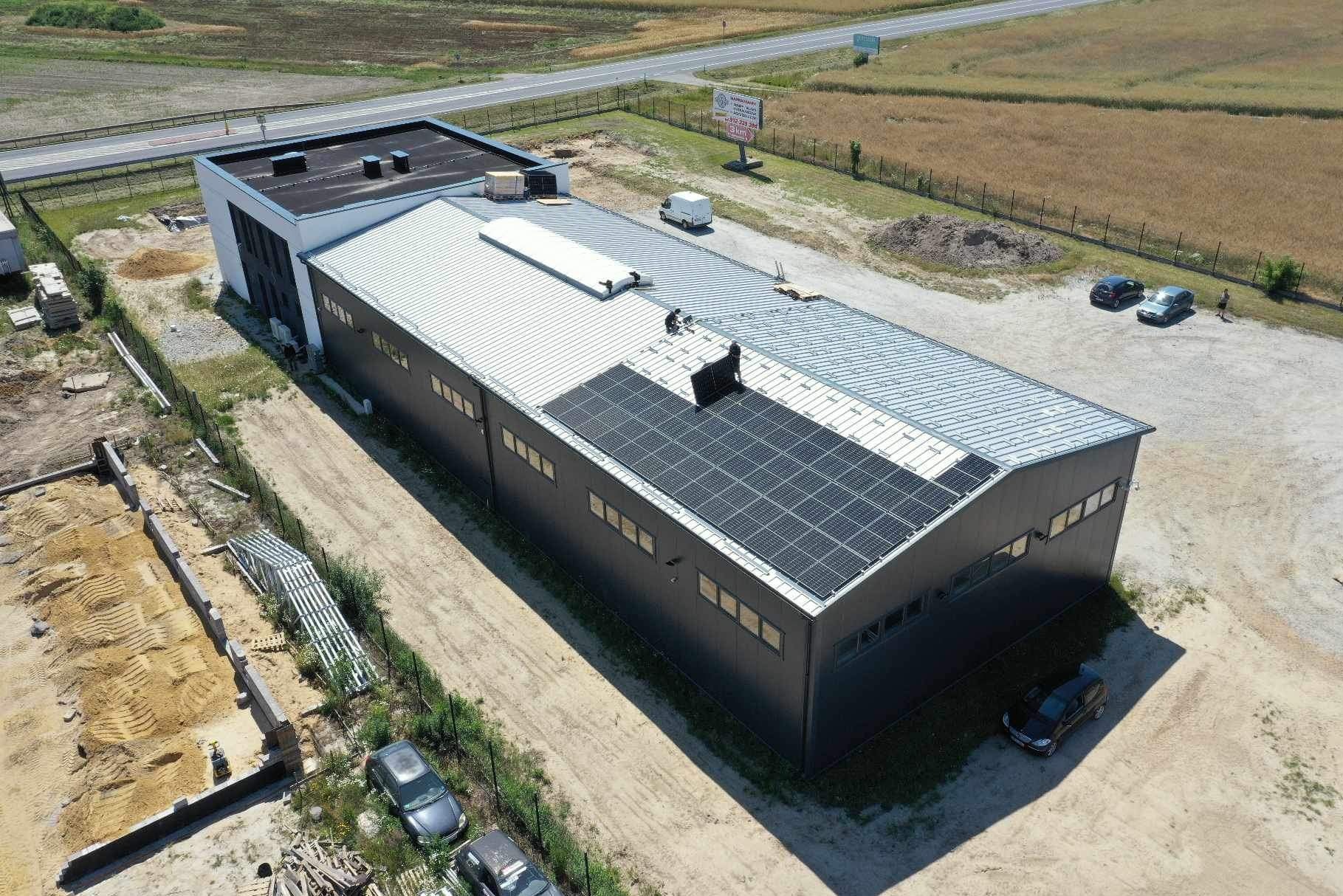 Widok z lotu ptaka na budynek przemysłowy z zamontowanymi panelami fotowoltaicznymi na dachu. Na dachu widoczni dwaj monterzy. Obok budynku plac z zaparkowanymi samochodami i elementami...