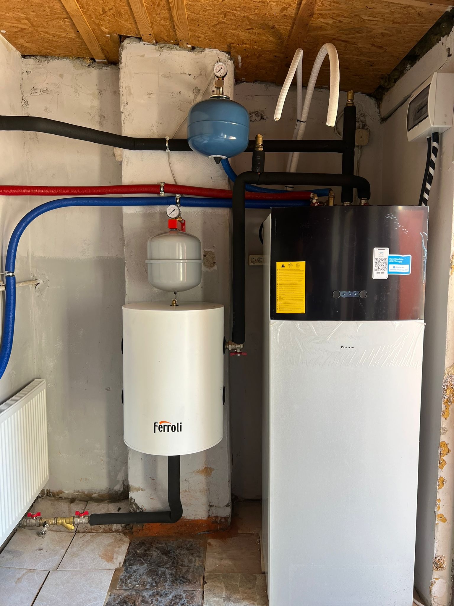 Instalacja grzewcza z białą pompą ciepła Daikin i zasobnikiem Ferroli, widoczne rury w izolacji, manometry, instalacja w pomieszczeniu z surowym wykończeniem ścian i podłogi.