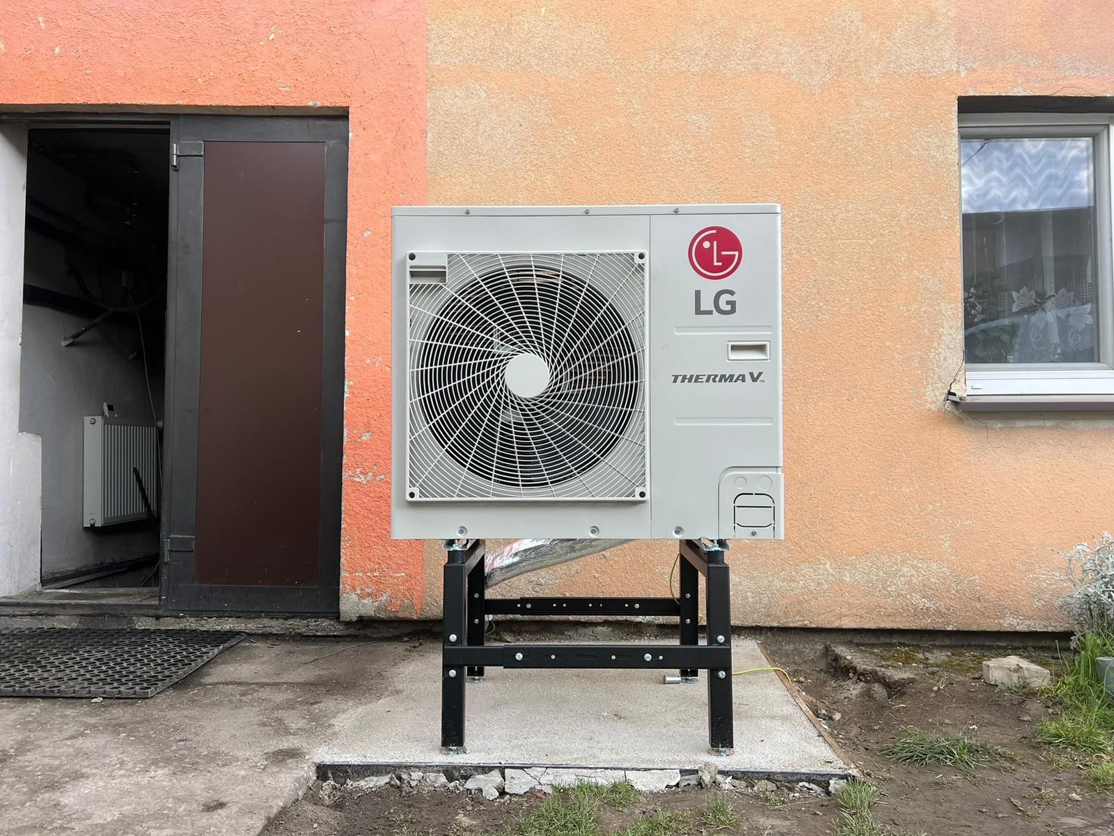 Zewnętrzna jednostka pompy ciepła LG Therma V na czarnej metalowej konstrukcji, ustawiona przed budynkiem z oknem i otwartymi drzwiami do piwnicy.