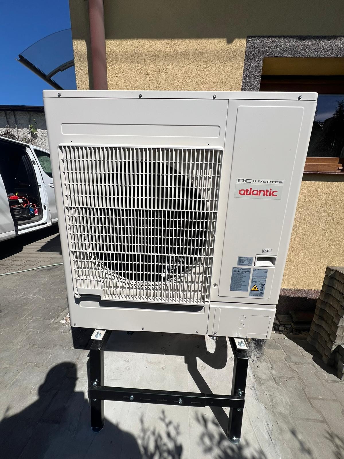 Kompaktowa, zewnętrzna jednostka pompy ciepła Atlantic DC Inverter, ustawiona na czarnym, metalowym stelażu przed budynkiem z żółtą elewacją, z widocznym samochodem dostawczym w tle.