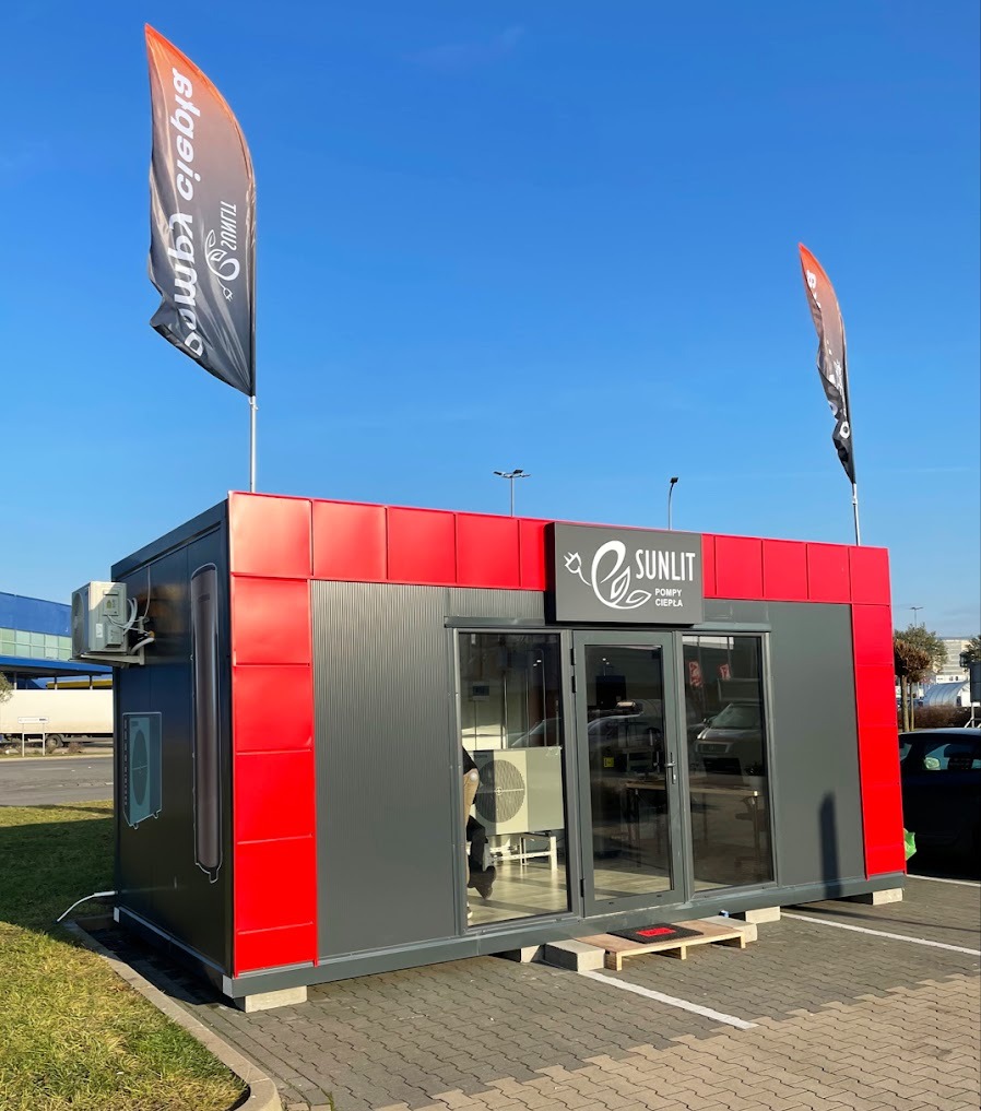 Mobilny pawilon demonstracyjny pomp ciepła Sunlit w Poznaniu, z widocznym logo i flagami reklamowymi na tle błękitnego nieba.