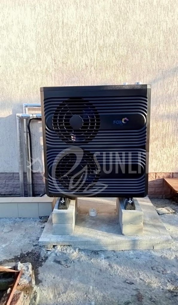 Czarna pompa ciepła marki FOX zamontowana na betonowych bloczkach przed budynkiem z rurami instalacyjnymi na ścianie.