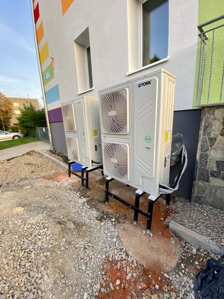 Dwie jednostki zewnętrzne pompy ciepła York zamontowane na czarnych, metalowych podstawach przed budynkiem z kolorową elewacją. Teren wokół w trakcie prac wykończeniowych, widoczne kamienie...