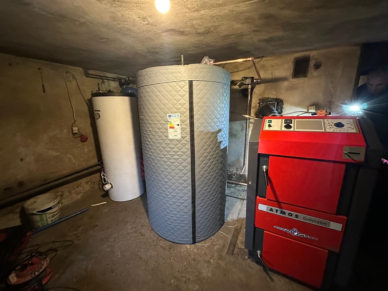 Piwnica z zainstalowanym piecem na paliwo stałe Atmos Generator, zbiornikiem akumulacyjnym z izolacją i bojlerem, oświetlana przez montera z latarką.