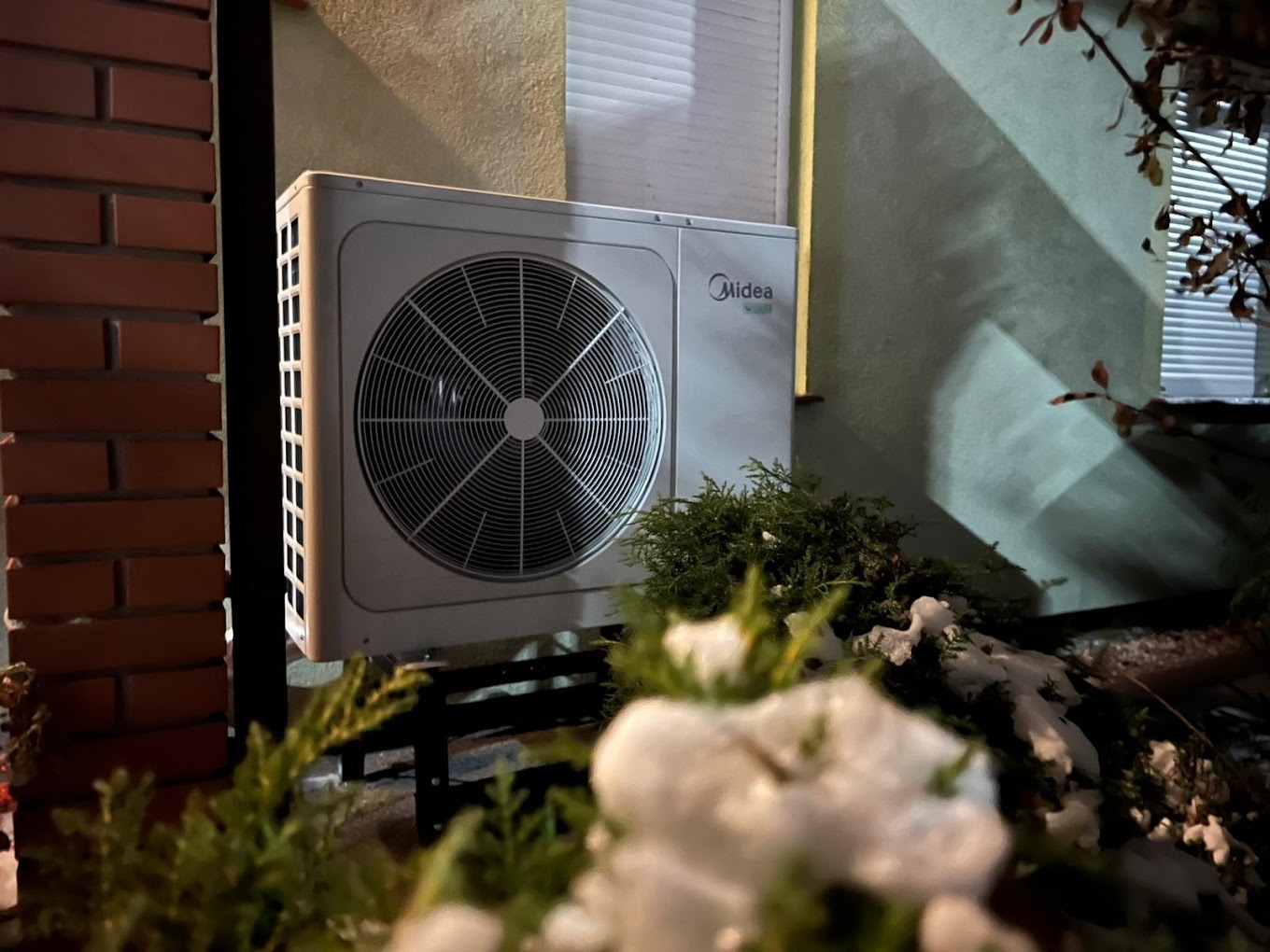 Zewnętrzna jednostka pompy ciepła Midea zamontowana na czarnej metalowej konstrukcji przy ścianie budynku z cegły i zielonym tynkiem, z widocznymi żaluzjami okiennymi i roślinnością w tle i...