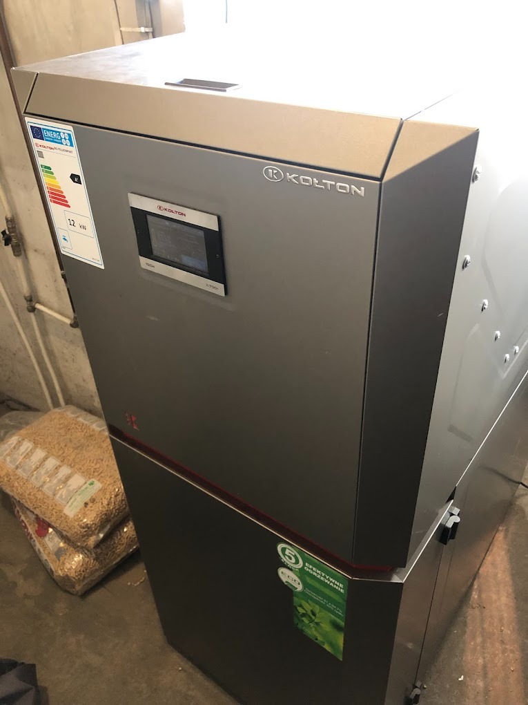 Szary piec na pellet marki Kolton z panelem sterowania, obok worki z pelletem, widoczne naklejki z klasą energetyczną i informacją o efektywnym ogrzewaniu.