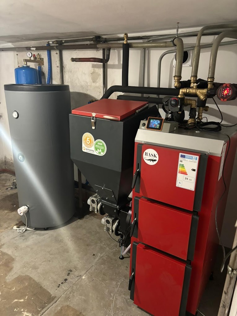 Szary zbiornik akumulacyjny ciepła obok czerwono-czarnego pieca na paliwo stałe z podajnikiem, widoczne naklejki ECO Design i logo producenta, instalacja w kotłowni.