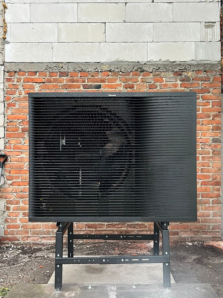 Czarna jednostka zewnętrzna pompy ciepła Daikin, zamontowana na metalowym stelażu przed ścianą z czerwonej cegły i białych bloczków.