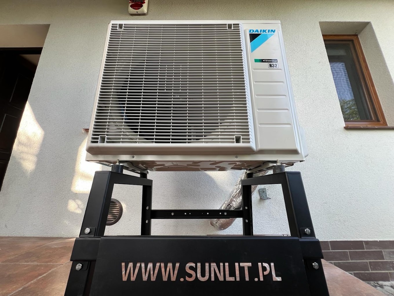 Biała jednostka zewnętrzna klimatyzacji Daikin Inverter R32 na czarnym, metalowym stojaku z adresem strony internetowej na tle jasnej elewacji budynku z oknem.