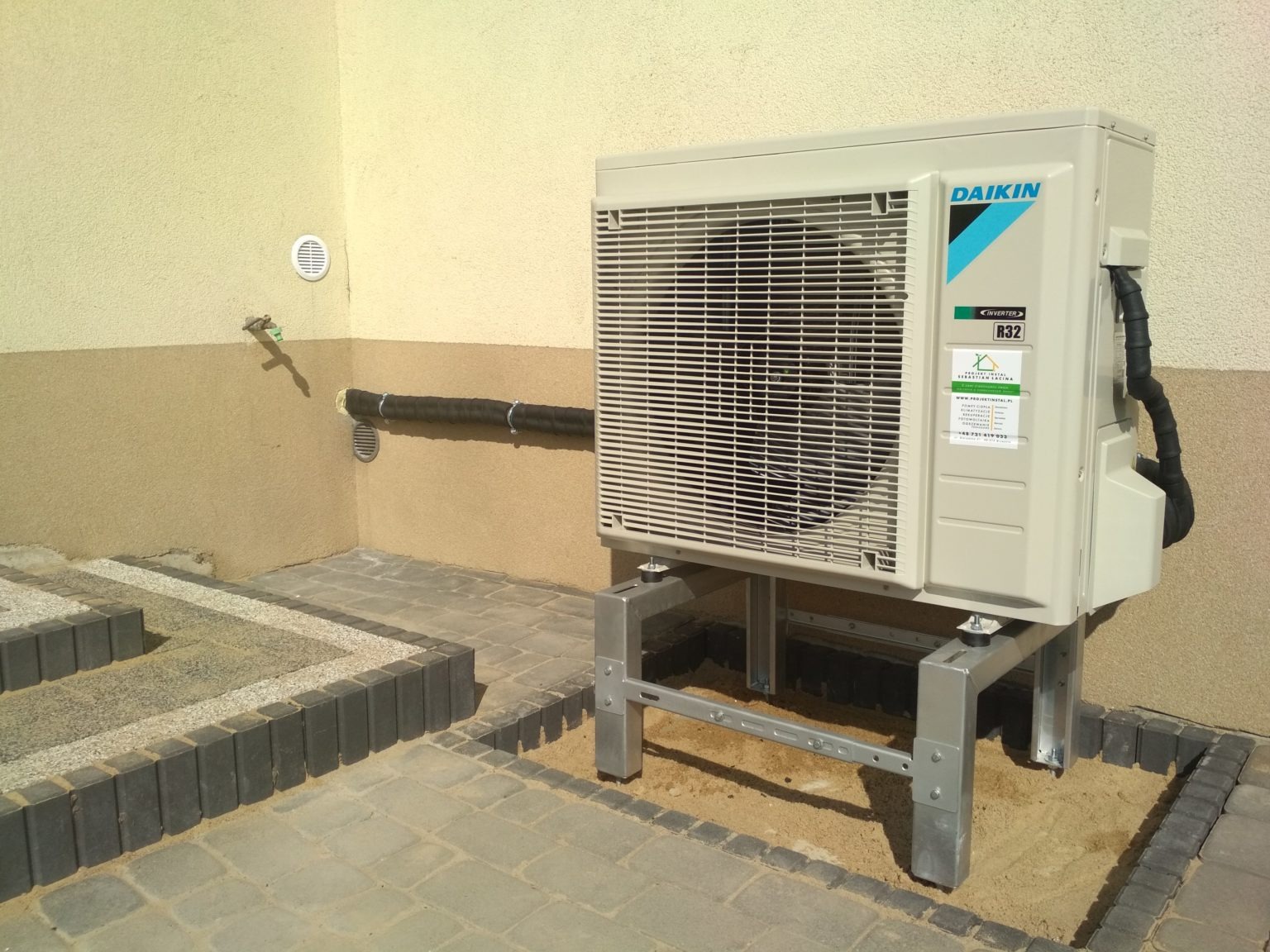 Zewnętrzna jednostka klimatyzacyjna Daikin na metalowej konstrukcji wsporczej, umieszczona na piasku otoczonym kostką brukową przy ścianie budynku. Widoczna rura zabezpieczona izolacją przymocowana...