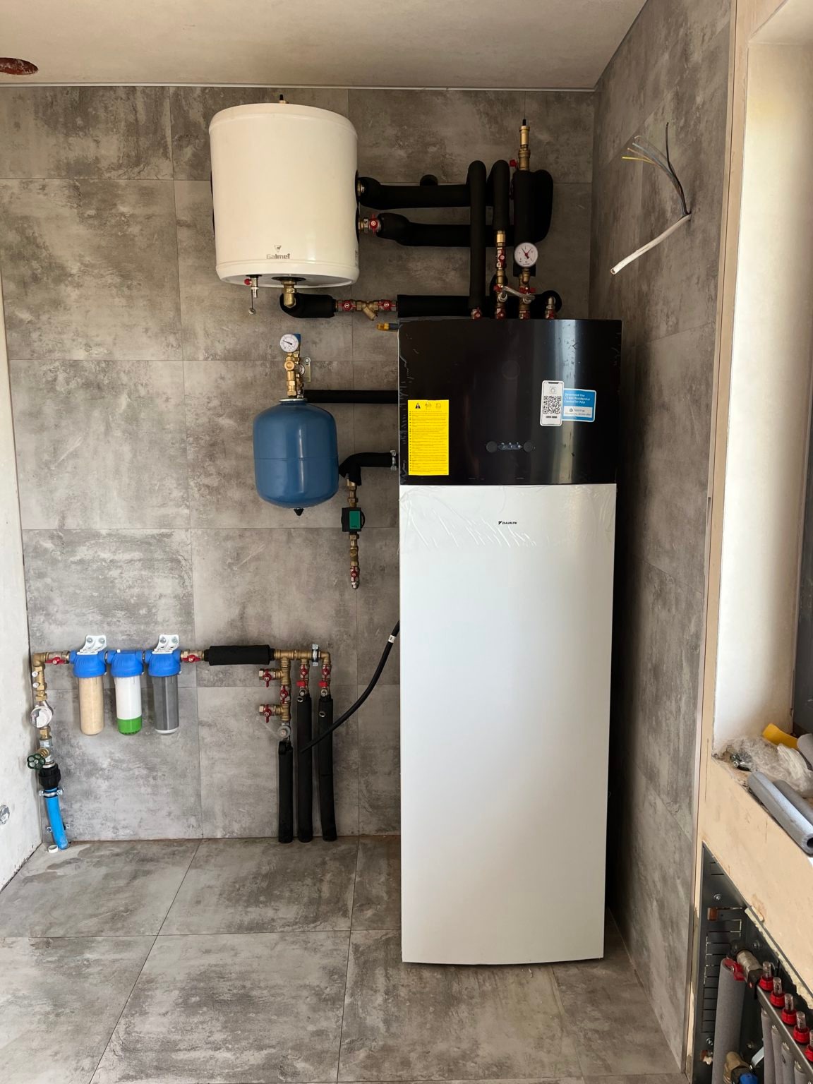 Biała pompa ciepła Daikin z czarnym panelem, zbiornik buforowy Galmet, filtry wody i rury miedziane na tle szarych płytek imitujących kamień.