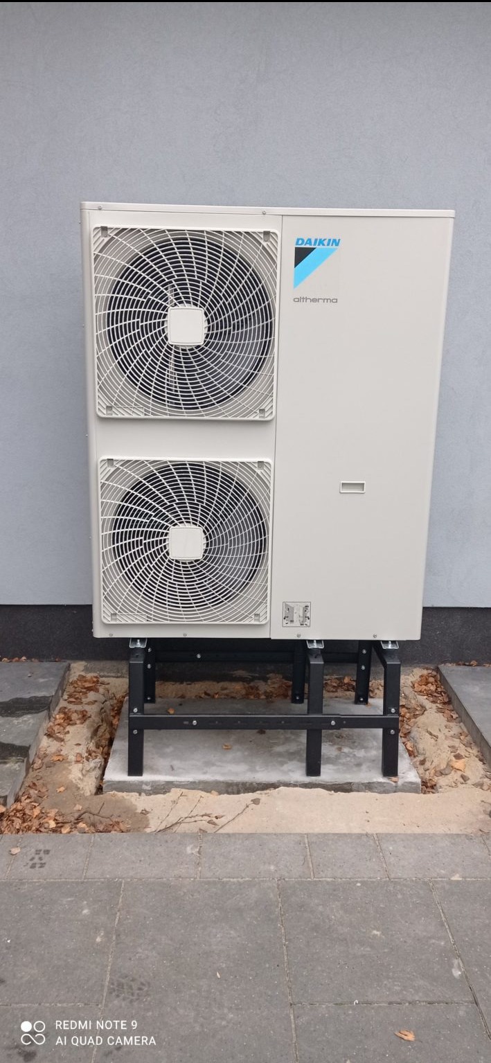 Zewnętrzna jednostka pompy ciepła Daikin Altherma, zamontowana na czarnej, metalowej konstrukcji wsporczej, ustawiona na betonowej podstawie obok chodnika z kostki brukowej i ściany budynku.