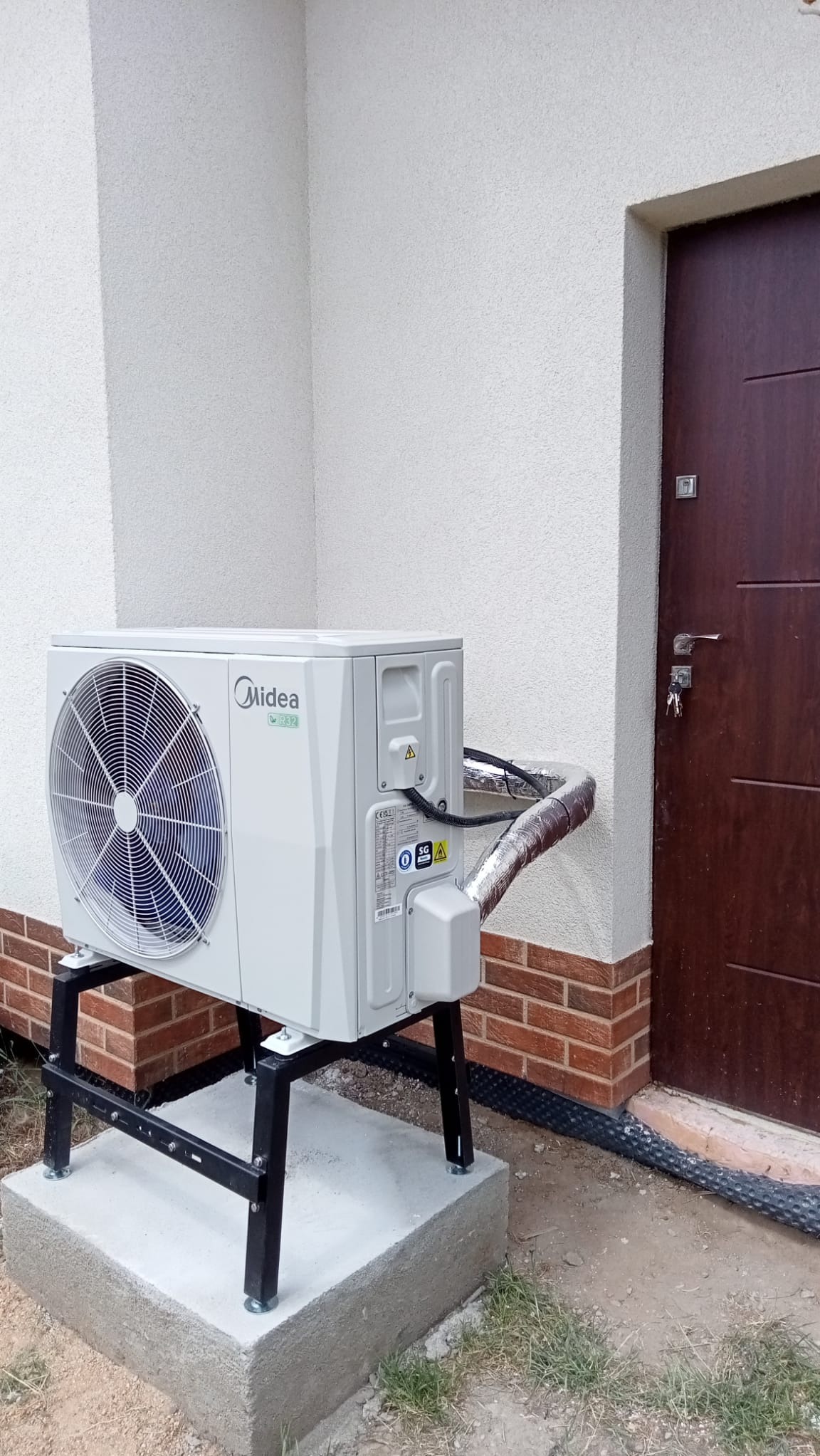 Biała jednostka zewnętrzna pompy ciepła Midea, zamontowana na czarnym metalowym stelażu na betonowej podstawie, z izolowanymi rurami przyłączonymi do ściany budynku obok brązowych drzwi wejściowych.