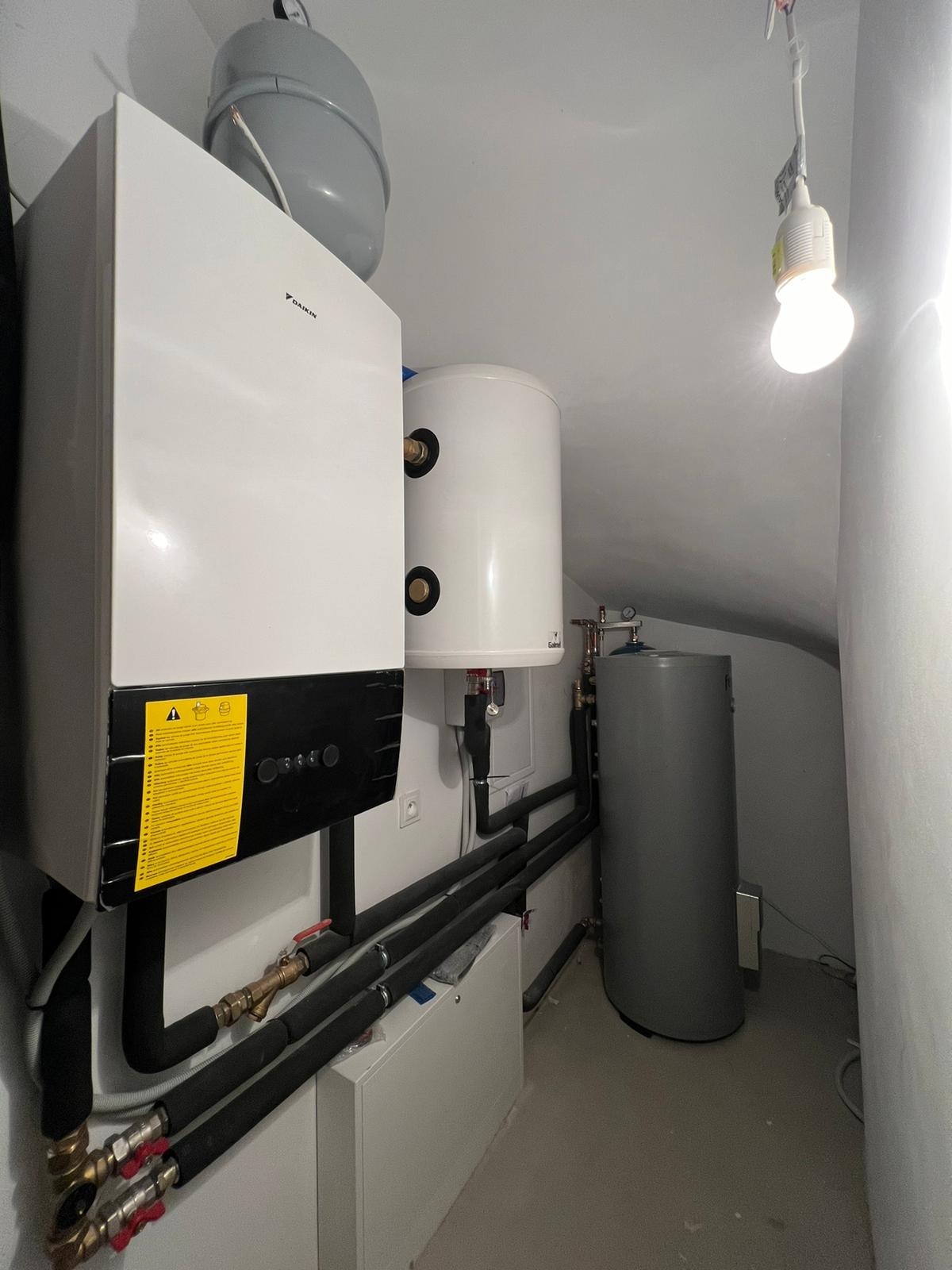 Zestaw urządzeń grzewczych w kotłowni: biały piec gazowy Daikin z żółtą naklejką informacyjną, biały zbiornik na wodę, szary zasobnik, czarne rury izolowane, oświetlenie żarówką na kablu.