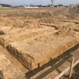 Wykopane rowy pod fundamenty na placu budowy, widoczny poziomica i drewniane szalunki, w tle pole i zabudowania.