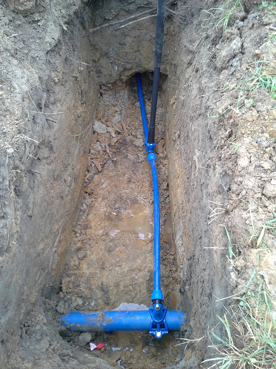 Wykop w ziemi z widocznymi niebieskimi rurami łączącymi się kształtkami, jedna czarna rura pionowo w górę, woda w wykopie.