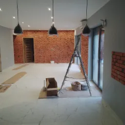 Wnętrze w trakcie wykańczania z ceglaną ścianą, aluminiową drabiną, marmurową podłogą, lampami wiszącymi w folii ochronnej i kartonem z narzędziami na podłodze.