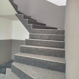 Patryk Jawor - Schody wykończone szarą, lastrykową okładziną z aluminiowymi listwami, prowadzące w górę. Ściany pomalowane w odcieniach szarości i bieli. Ujęcie z dołu.