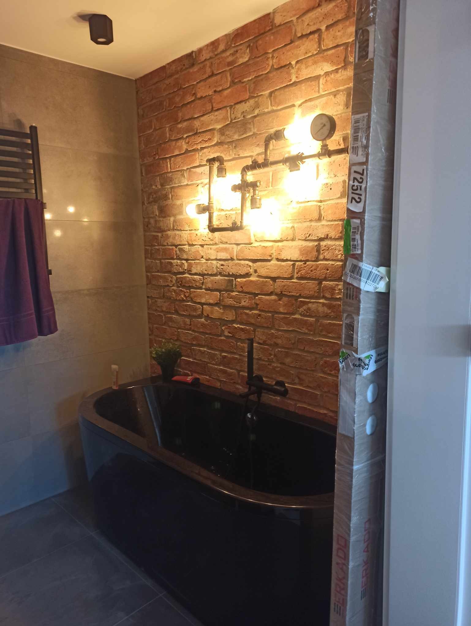 Stylowa łazienka z czarną wanną na tle ceglanej ściany z oświetleniem z rur. Elementy dekoracyjne i nowoczesny design. Widoczny fragment drzwi z opakowaniem.