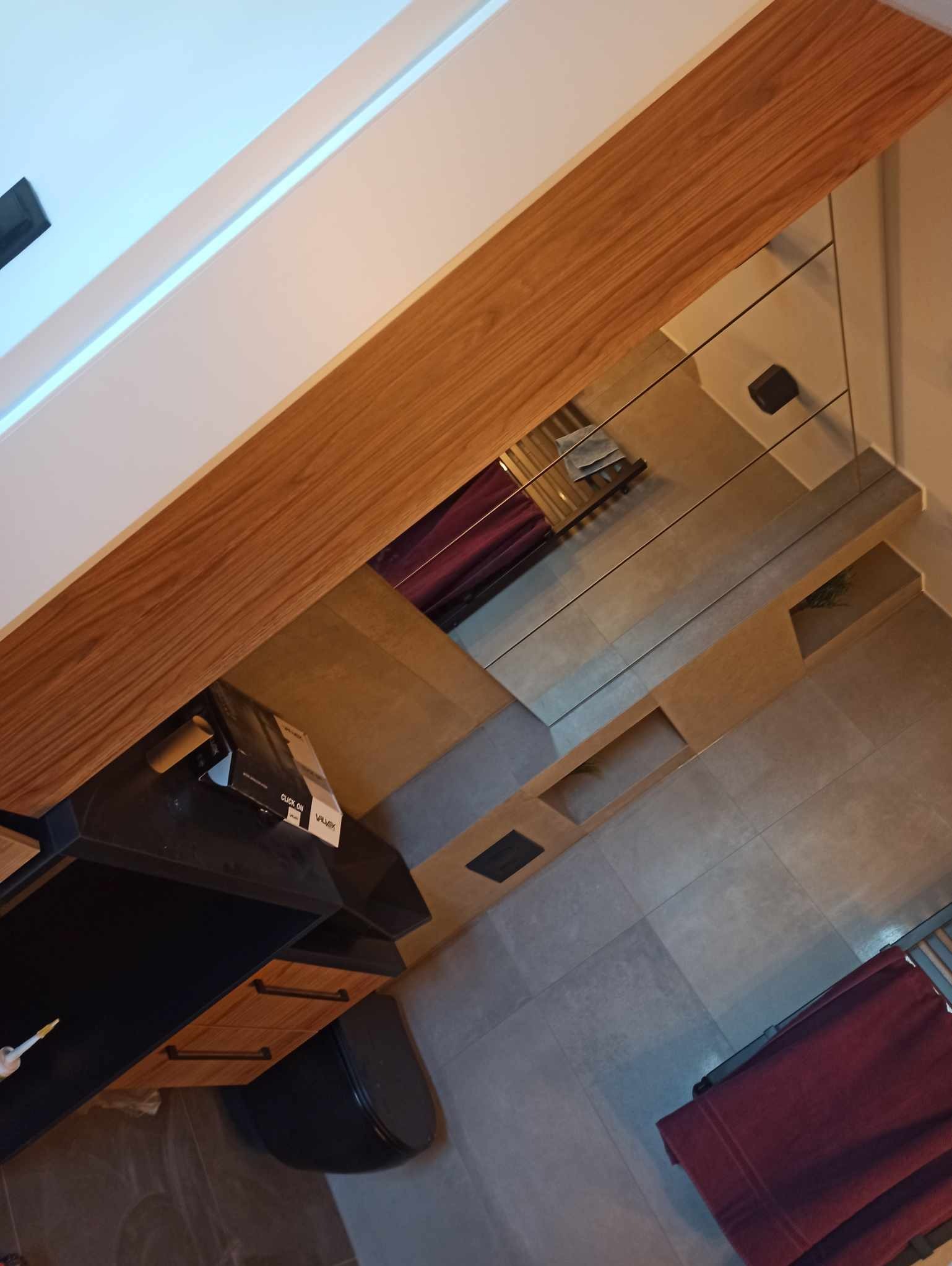 Nowoczesna łazienka z drewnianym blatem, lustrem i szarą podłogą. Uwagę przykuwa wisząca toaleta i minimalistyczny design. Widoczny fragment czerwonego ręcznika.