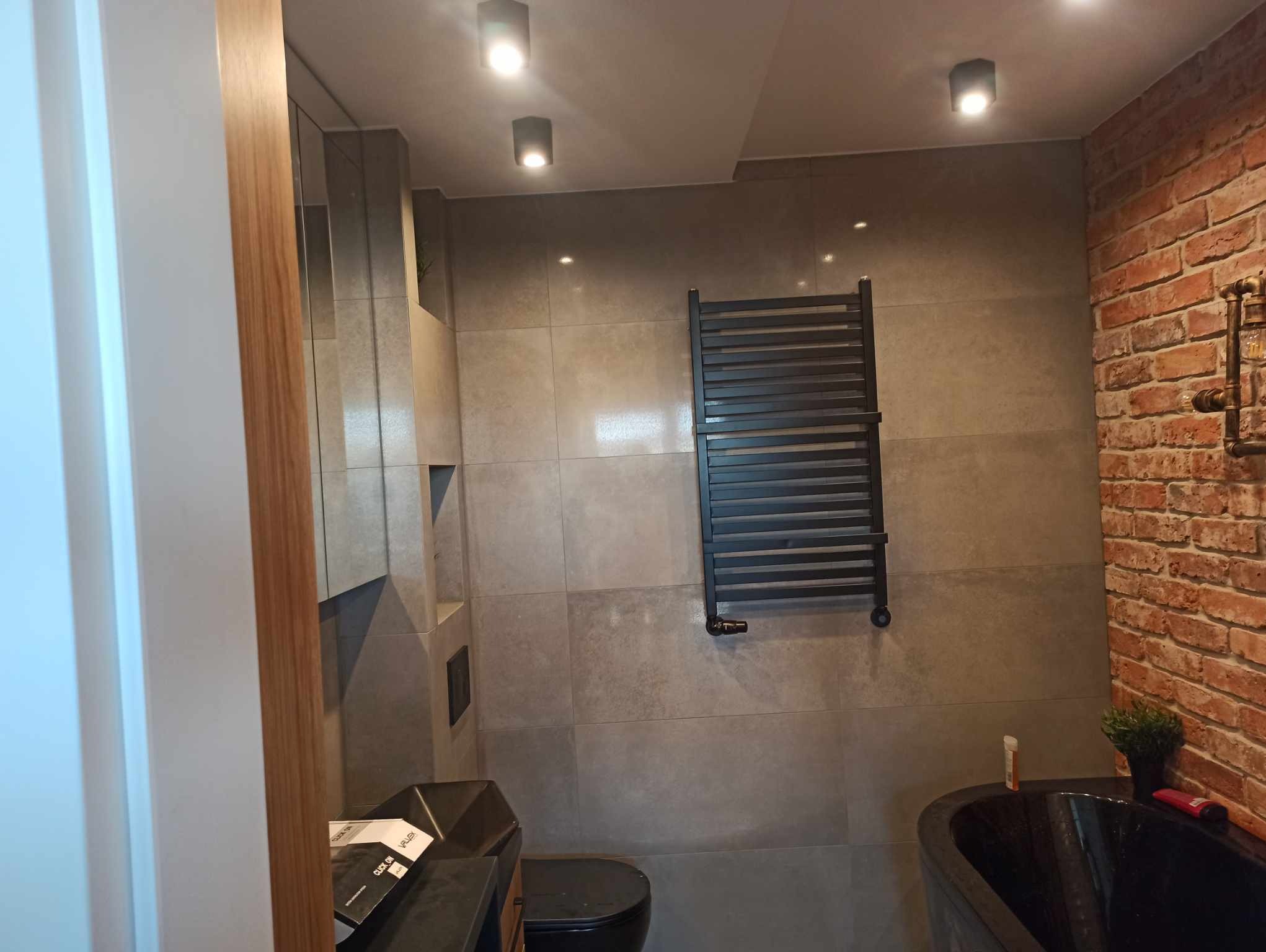 Nowoczesna łazienka z szarymi płytkami, ceglaną ścianą i czarną armaturą. Grzejnik drabinkowy i designerska umywalka dodają elegancji. Oświetlenie punktowe.