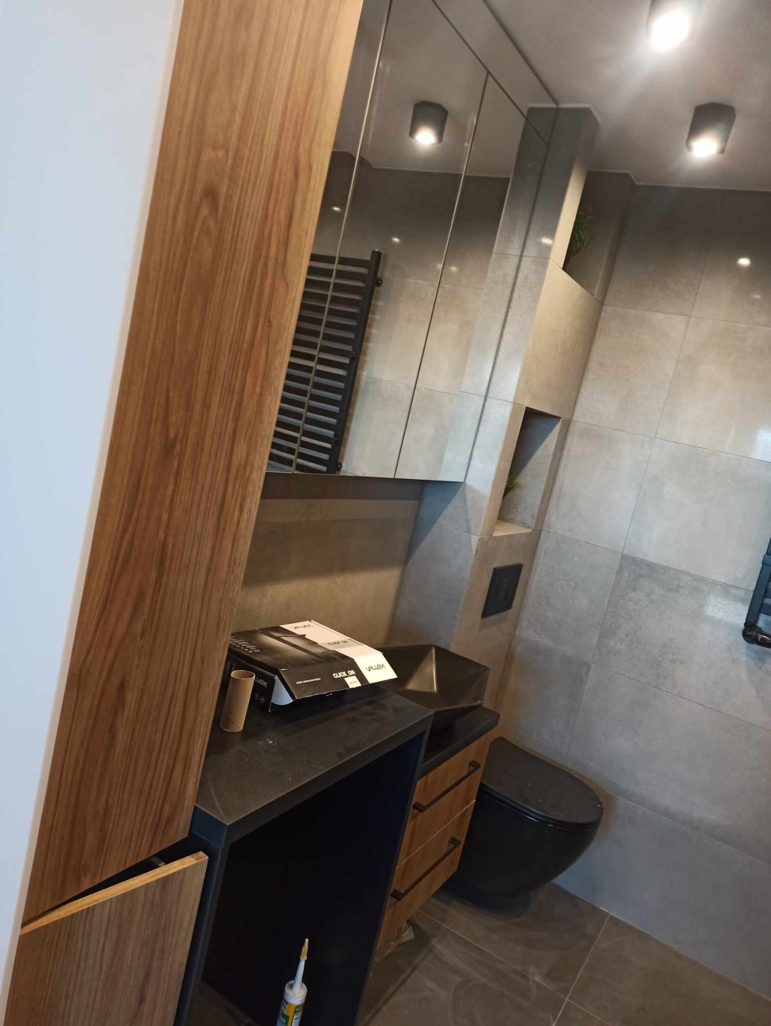 Nowoczesna łazienka z szarymi płytkami, czarną armaturą i drewnianymi elementami wykończeniowymi. Widoczna umywalka, toaleta i grzejnik.