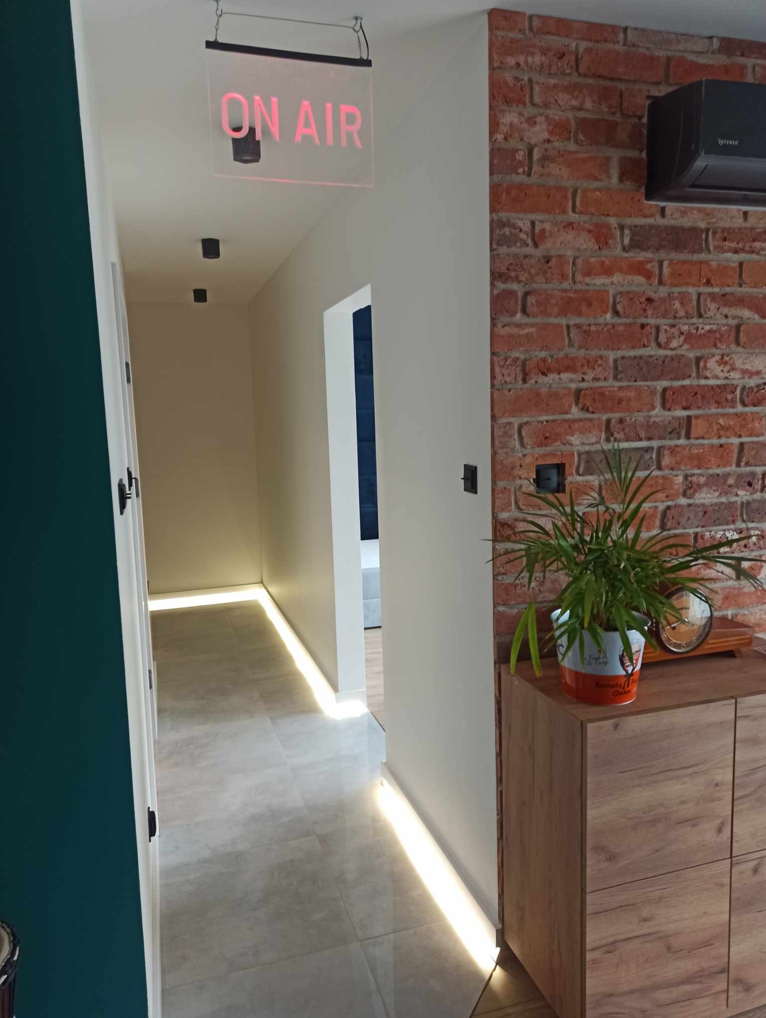 Korytarz po remoncie z oświetleniem podłogowym LED, ceglaną ścianą i wiszącą lampą 'ON AIR'. Widok na odnowione wnętrze z nowoczesnym designem.