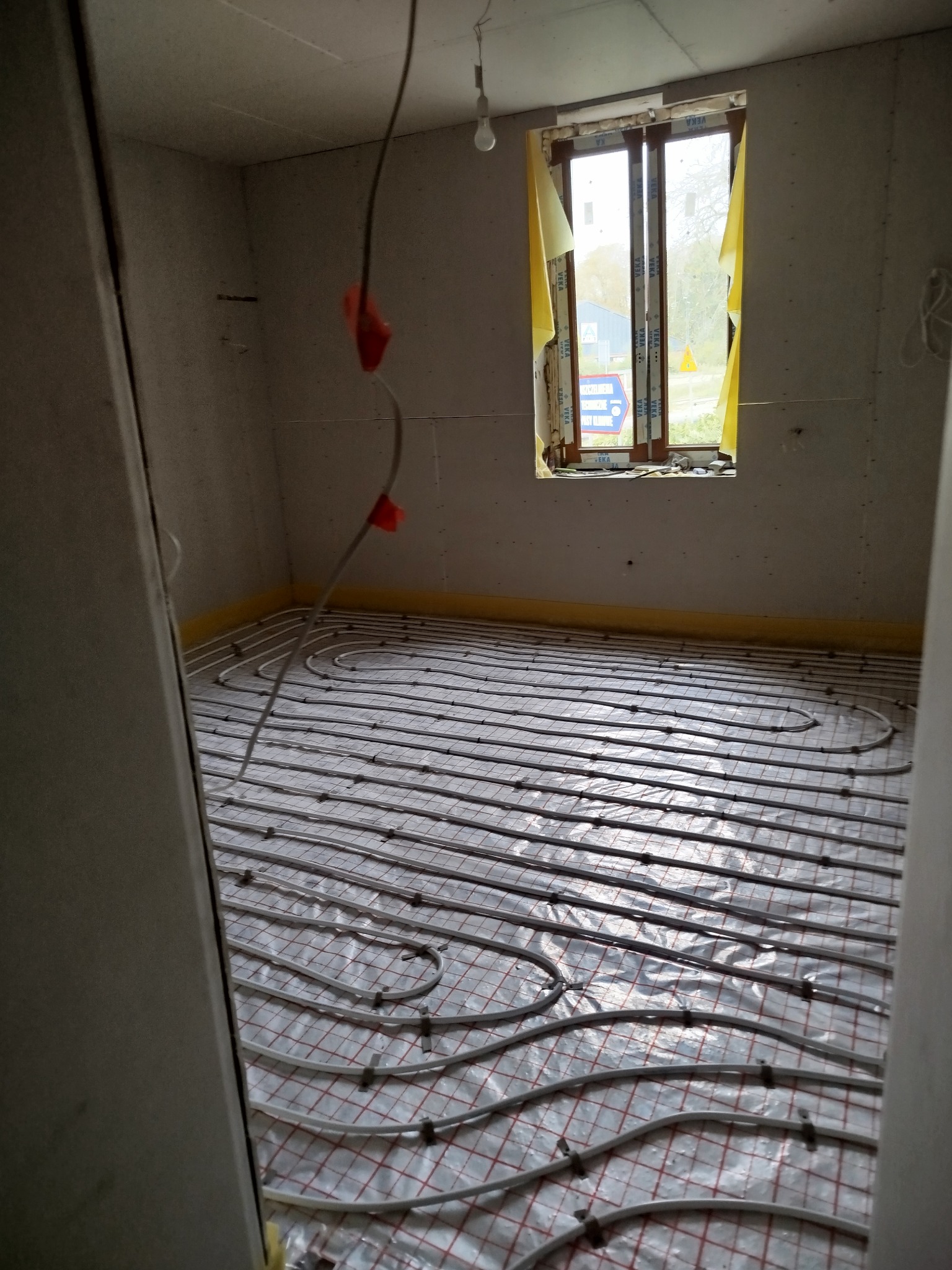 Instalacja ogrzewania podłogowego w surowym wnętrzu z płytami kartonowo-gipsowymi i oknem z żółtą folią ochronną. Widok na ułożone rury grzewcze.