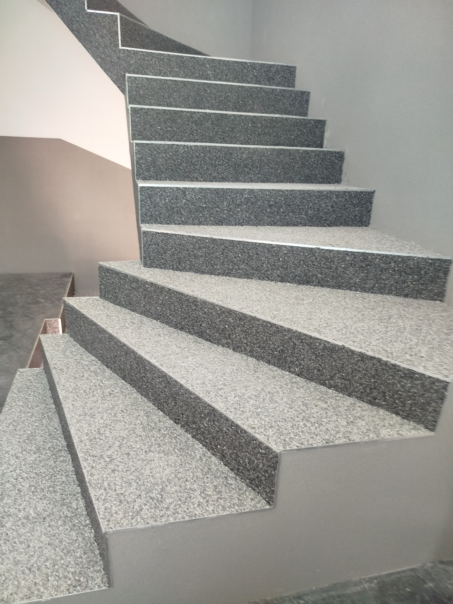 Schody wewnętrzne wykończone szarą, kamyczkową okładziną, widoczne z dołu pod kątem, z fragmentem ściany w tle. Detal wykończenia wnętrza.