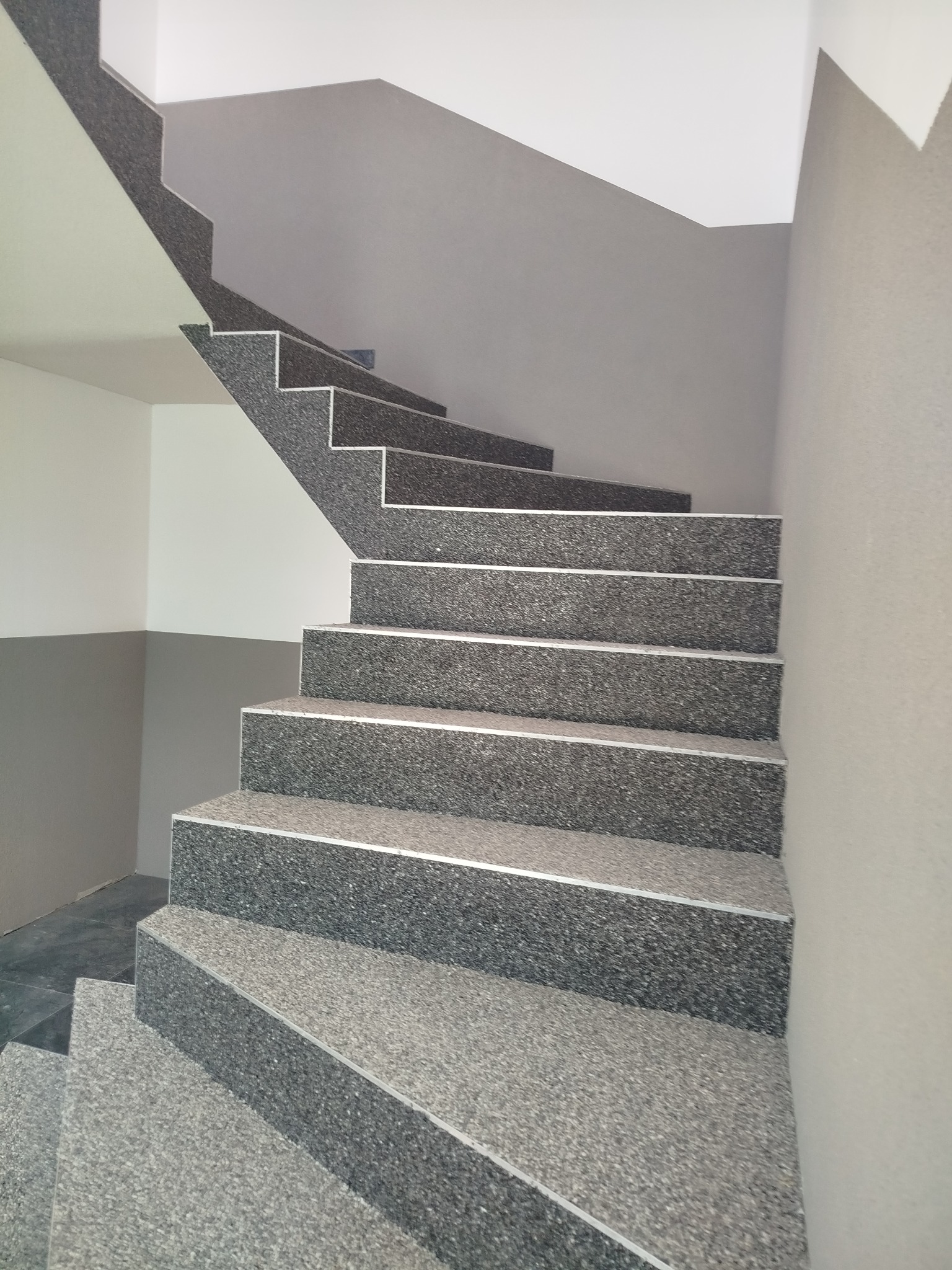 Schody wykończone szarą, lastrykową okładziną z aluminiowymi listwami, prowadzące w górę. Ściany pomalowane w odcieniach szarości i bieli. Ujęcie z dołu.