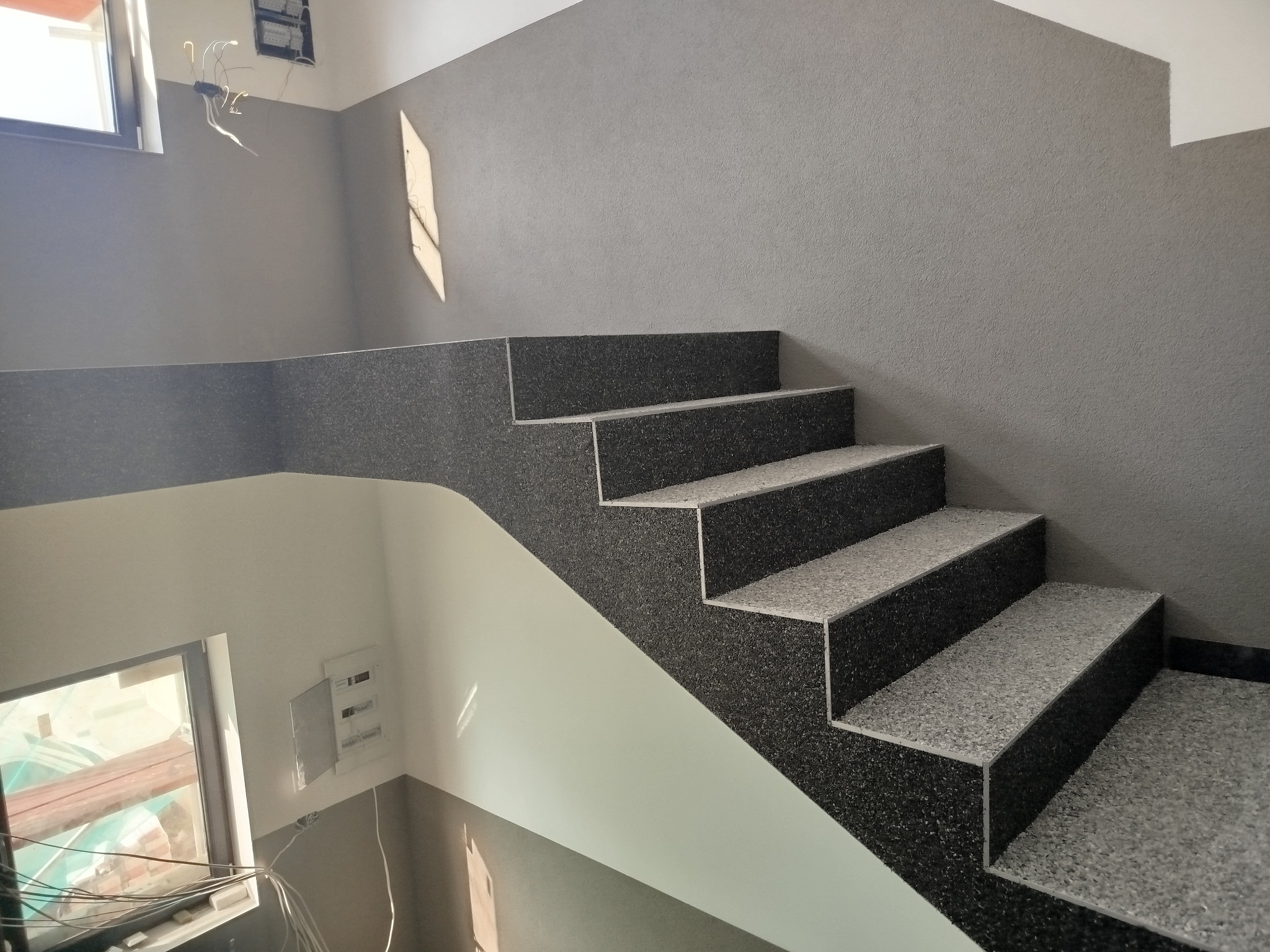 Wykończone schody wewnątrz budynku, pokryte szarą, wzorzystą okładziną. Widoczne okno i instalacja elektryczna. Ściany w odcieniach szarości i bieli. Ujęcie z dołu.