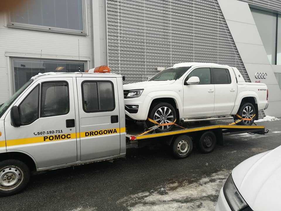 Biały pickup Volkswagen Amarok zabezpieczony pasami na platformie lawety pomocy drogowej przed salonem Audi w pochmurny dzień.