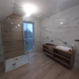 Łazienka w trakcie wykańczania: kabina prysznicowa ze szkła, drewnopodobne płytki podłogowe i ścienne, umywalka nablatowa na drewnianej szafce bez frontów, widoczne wystające kable.