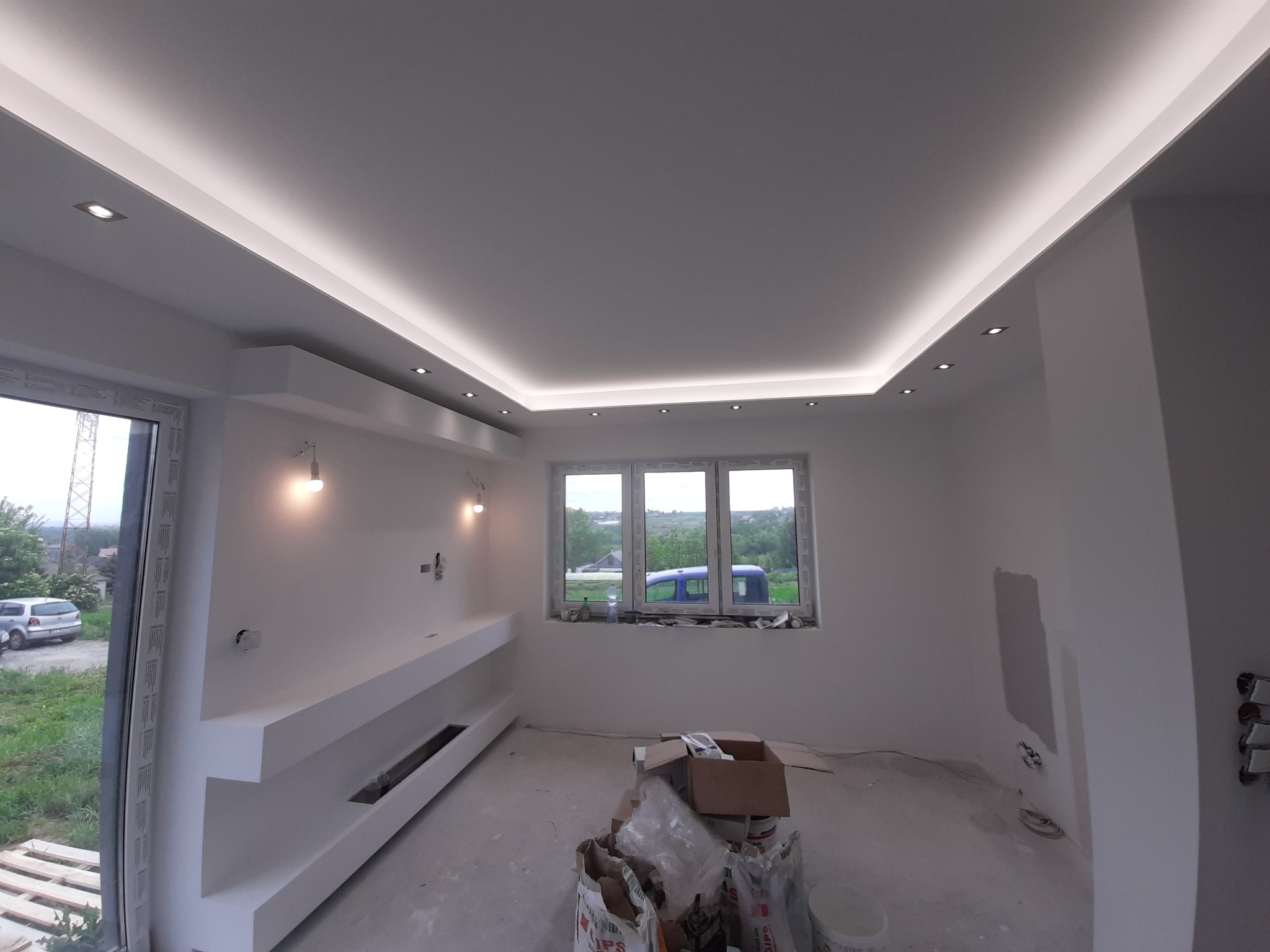 Wnętrze pokoju w trakcie wykończenia: białe ściany, podwieszany sufit z oświetleniem LED, wnęki na ścianie, okno z widokiem na zieleń, worki z materiałami budowlanymi na podłodze.