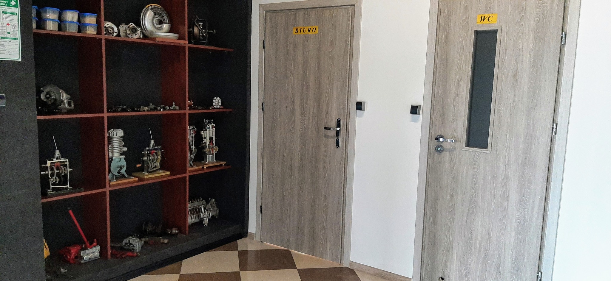 Wnętrze szkoły jazdy: regał z modelami silników i przekładni, obok drzwi do biura i toalety.
