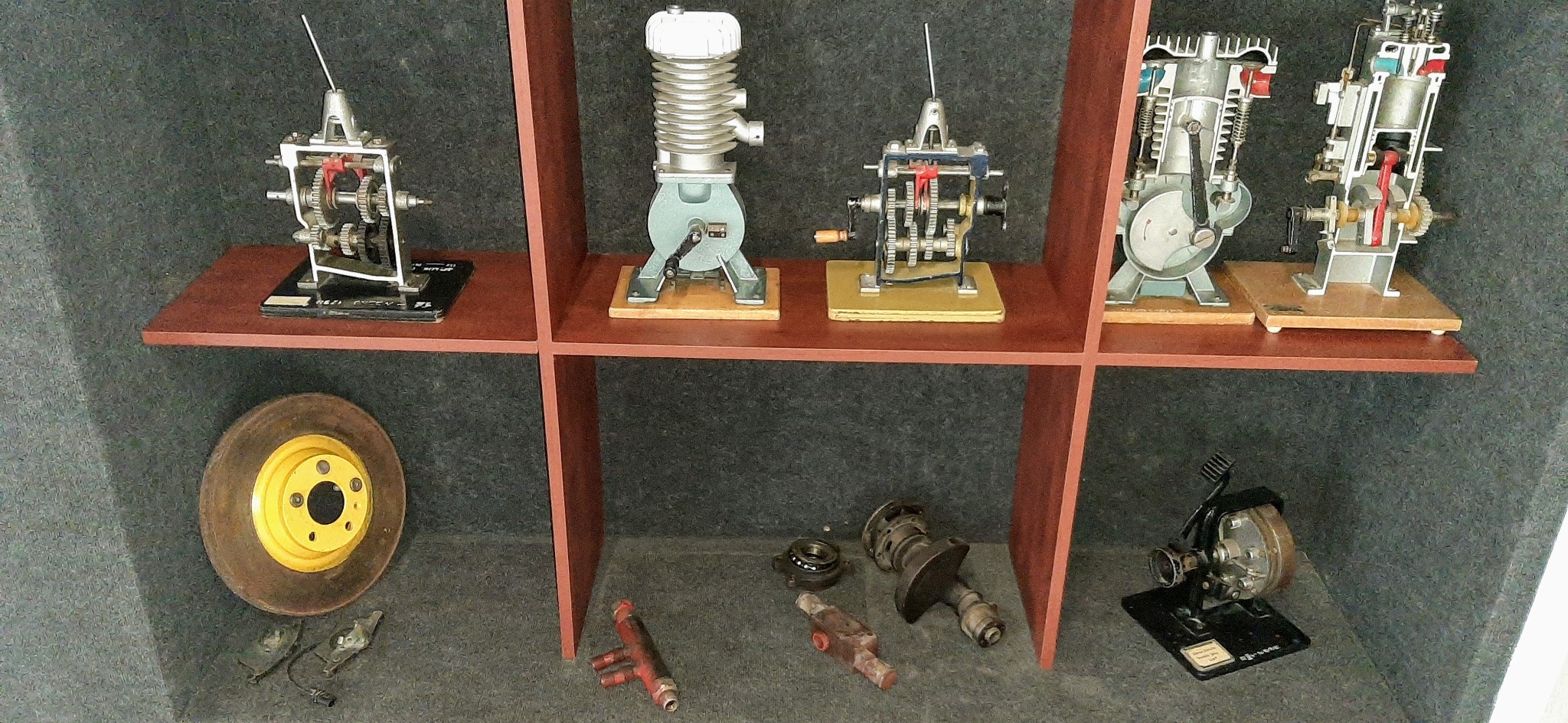 Modele edukacyjne silników spalinowych i przekładni na półkach, widoczny przekrój cylindra i tłoka, koło samochodowe z żółtą felgą i elementy zawieszenia.