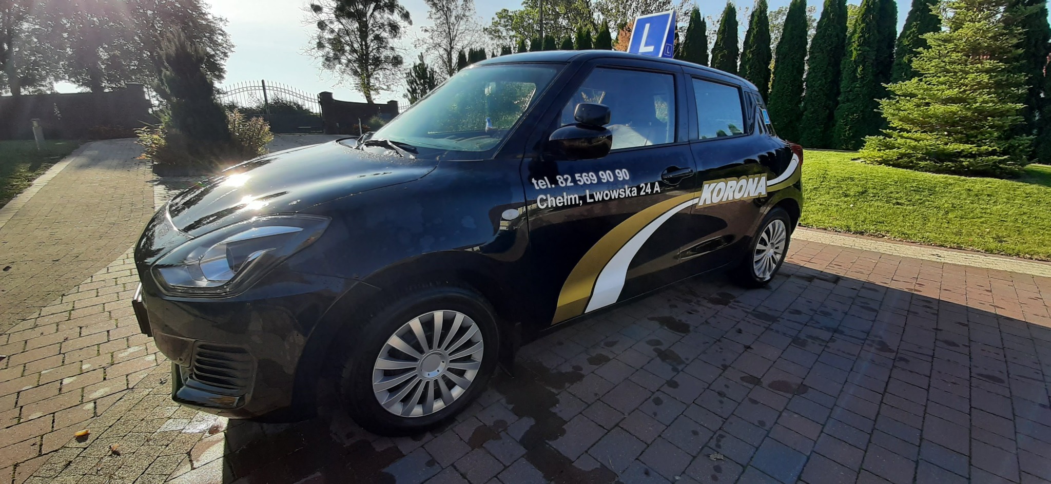 Czarny Suzuki Swift Hybrid z naklejkami i oznaczeniem 'L' na dachu, zaparkowany na brukowanej nawierzchni przed zielonymi tujami.