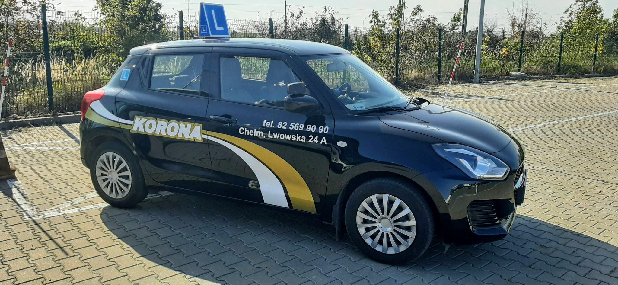 Czarny Suzuki Swift Hybrid z oznaczeniem 'L' na dachu i naklejkami z nazwą i numerem telefonu firmy, zaparkowany na placu z kostki brukowej.