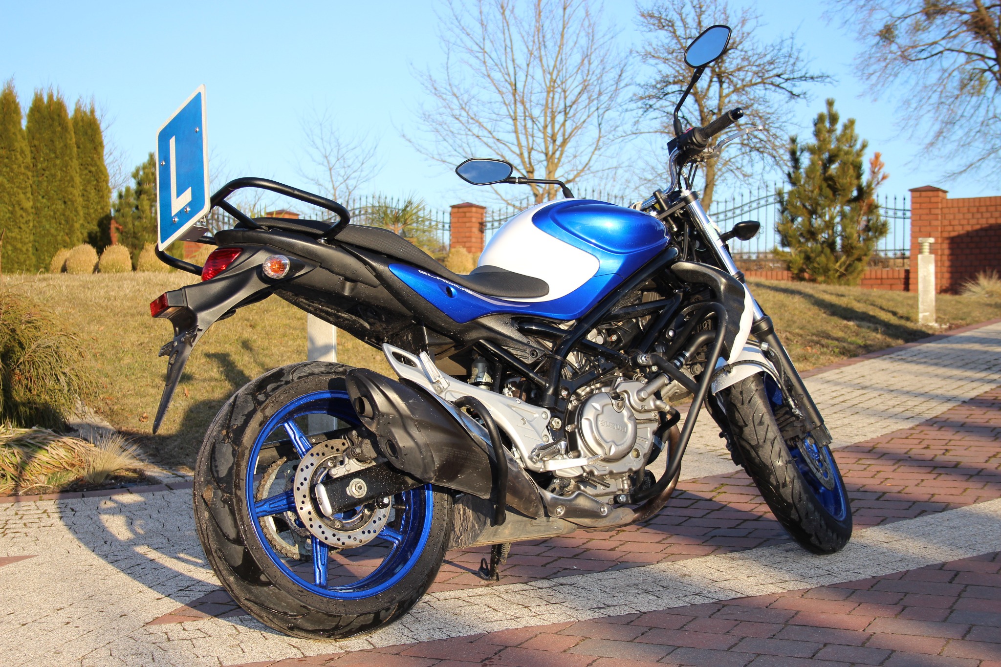Niebiesko-biały motocykl z tablicą 'L' na bagażniku, zaparkowany na chodniku z kostki, w tle trawnik i iglaki.