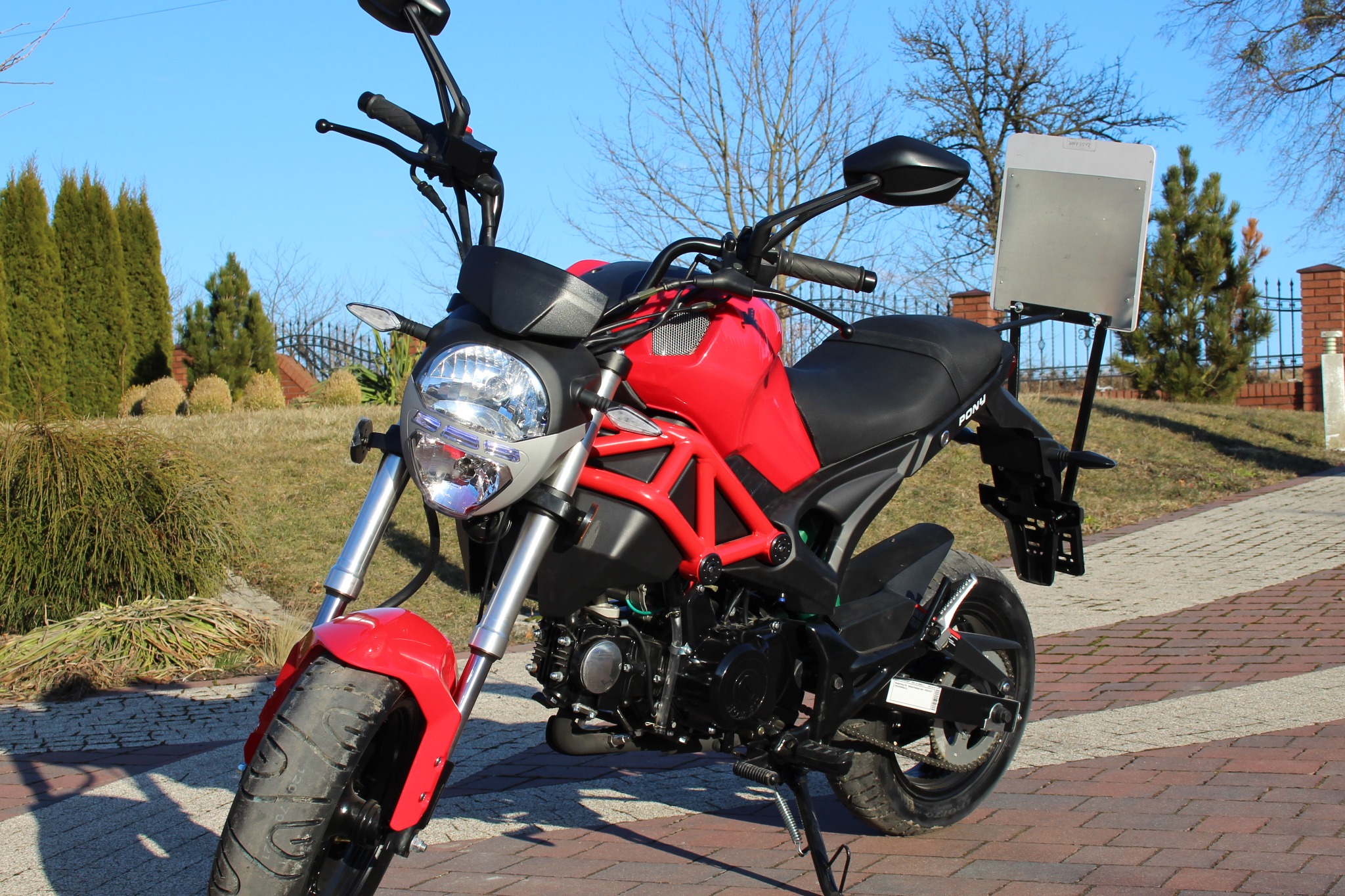 Czerwony motocykl z tablicą do nauki jazdy na tylnym błotniku, zaparkowany na kostce brukowej w słoneczny dzień.